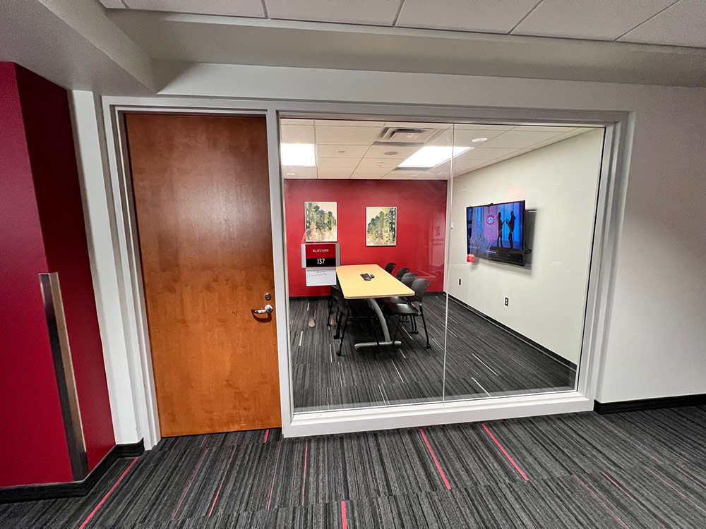View through the glass wall into the Blizzard Room at Atwood Memorial Center showing a table with chairs and a large computer monitor on a white wall
