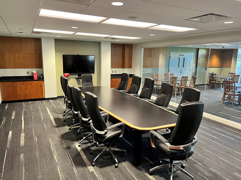Monarch Room interior at Atwood Memorial Center from a side angle showing the long conference table with black leather chairs, a wall‑mounted screen, wood cabinetry, and windows into an adjacent seating area