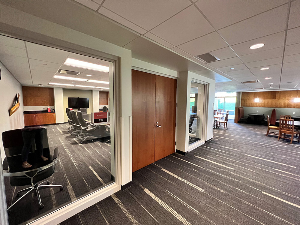 View from outside the Monarch Room in Atwood Memorial Center looking through glass into the room, with the conference table and chairs visible and a hallway seating area beyond