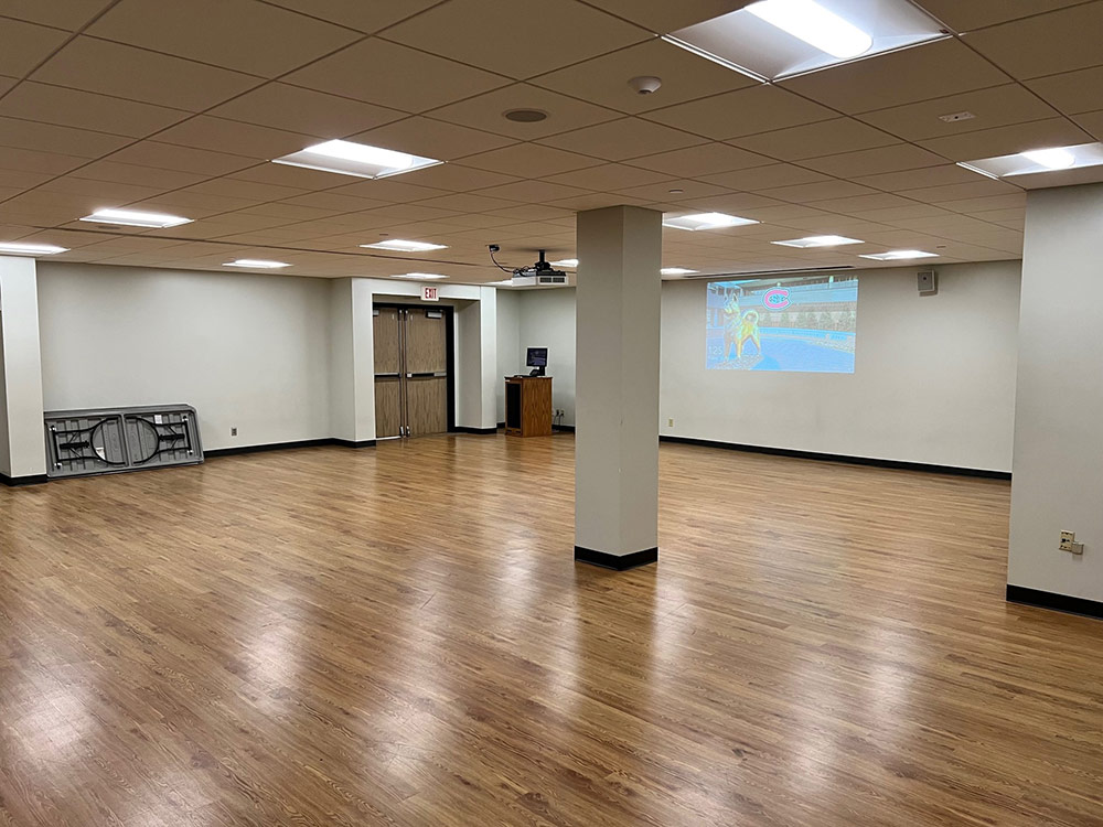 Interior of Atwood Memorial Center rehearsal room from a different angle showing hardwood floor, columns, a desktop screen, and folded tables against a wall