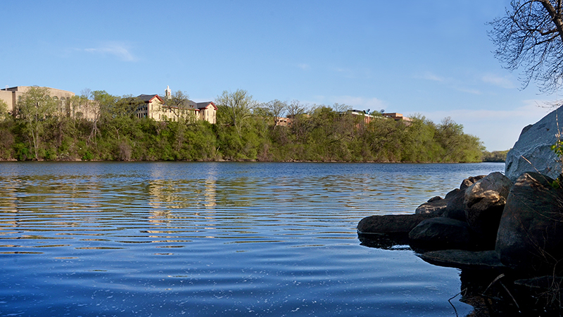 View of the Mississippi River with the St. Cloud State University campus in the background and trees along the riverbank on a sunny day