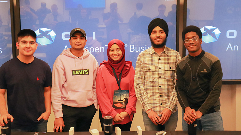 Five students stand together for a group photo, with three large screens visible in the background