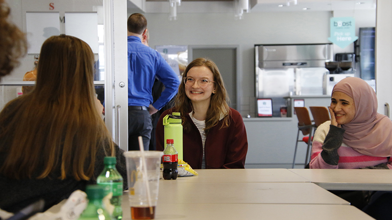 Three students sit at a table with beverages, engaged in a group discussion