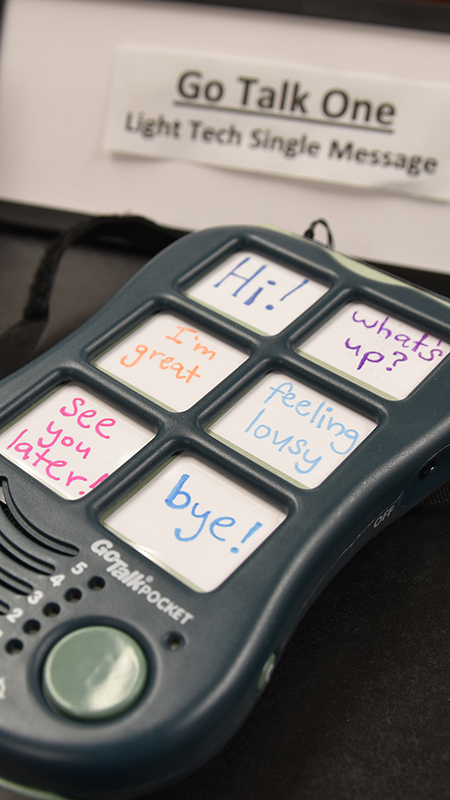 A handheld speech-generating device with large labeled buttons showing common phrases rests on a table