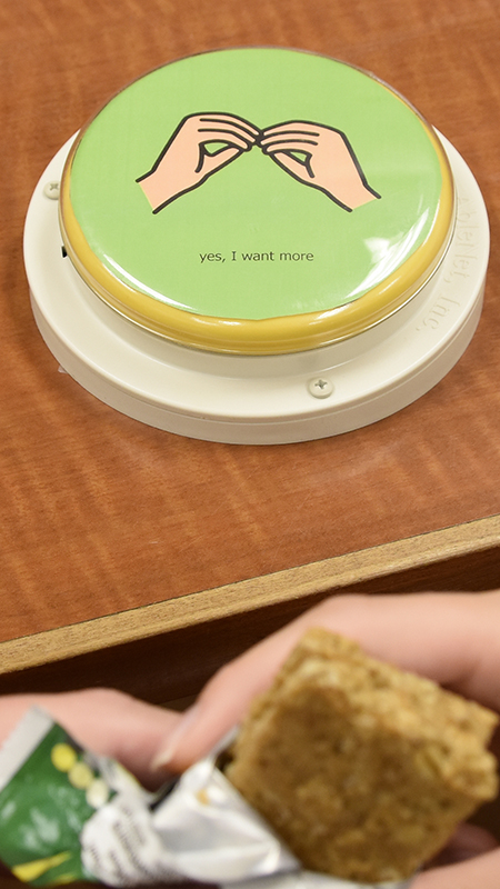 A large round communication button sits on a tabletop, showing a hand symbol and the words 'yes, I want more'