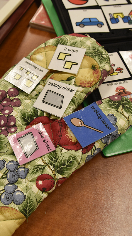 Oven mitts covered with picture-based instruction stickers lie on a table next to a communication board with illustrated symbols