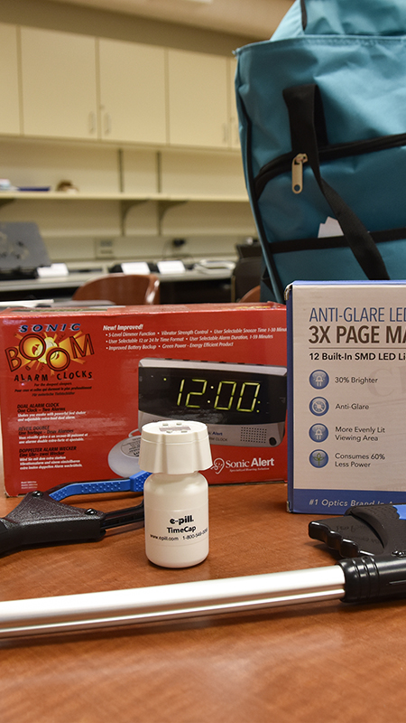 Assistive devices including a page magnifier, talking alarm clock, pill organizer, and other tools are arranged on a table in an indoor setting