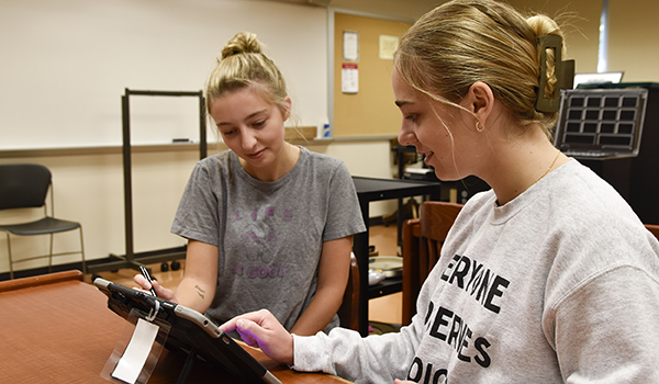 Two students use a tablet together in a classroom, with a large whiteboard visible in the background