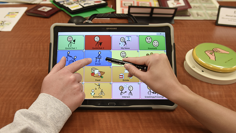 Two hands interact with a touchscreen tablet displaying a grid of illustrated communication icons on a table, with a large push button device nearby