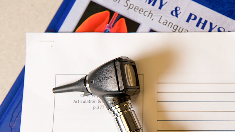 A handheld hearing examination instrument rests on paperwork and books related to speech and hearing studies