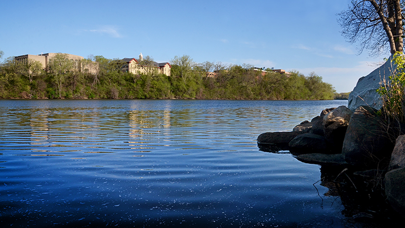 View of the Mississippi River with the St. Cloud State University campus in the background and trees along the riverbank on a sunny day