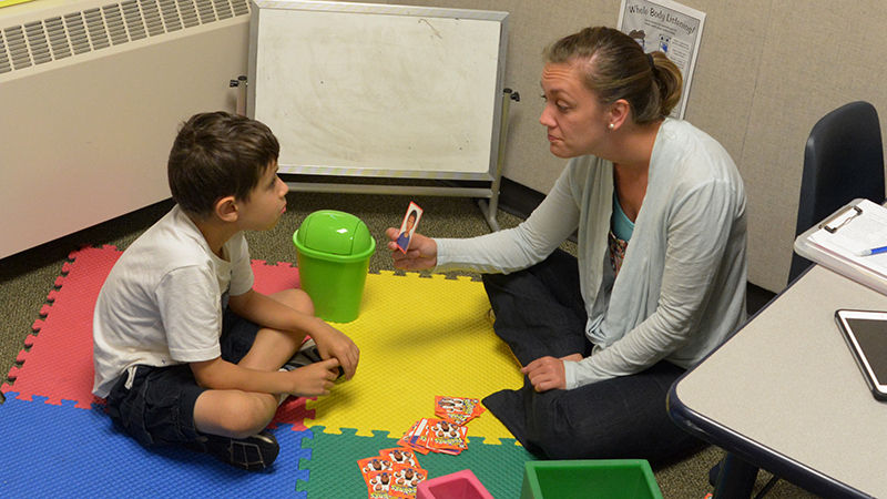 An instructor holds up a picture while working with a student seated on a colorful mat, with toys visible nearby
