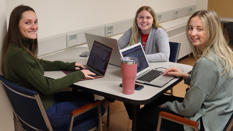 Three students work together on laptops at a table in a study area