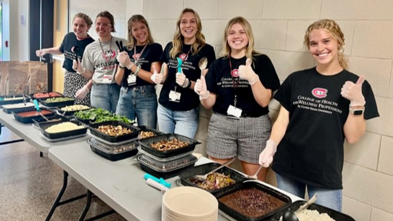 Six students stand together behind a table displaying various food dishes prepared for an event
