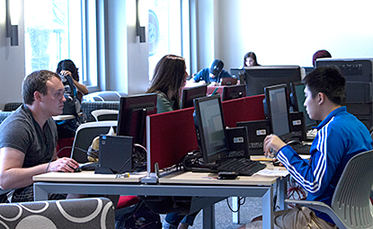 Several students sit at computer workstations in a computer lab, working on coursework at desktop computers