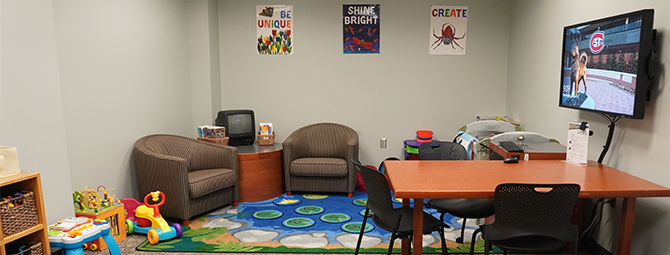 Interior view of a student parent study room with tables, chairs, a wall‑mounted computer screen, and children's toys visible throughout the space