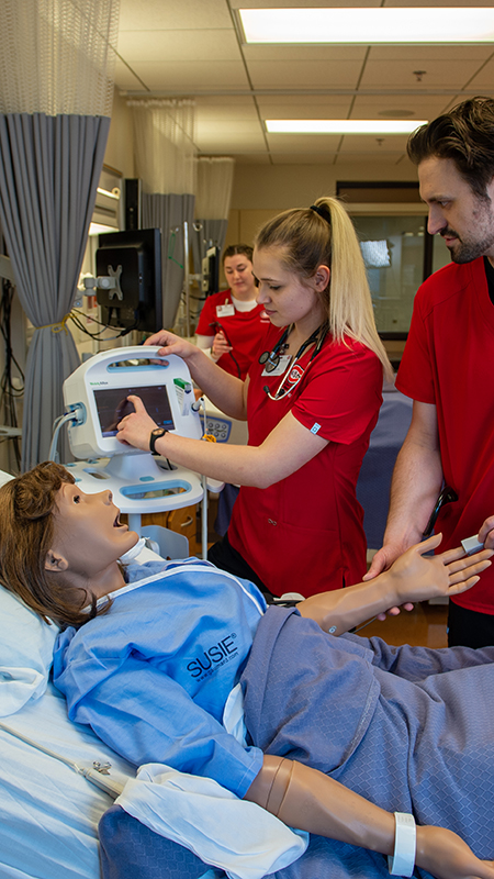 Nursing professor making a hand gesture while speaking to three students in a simulation lab