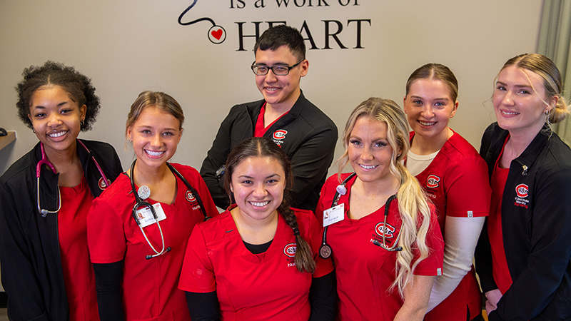 Nursing students standing in front of a wall with the phrase 'Nursing is a work of heart' for a group photo