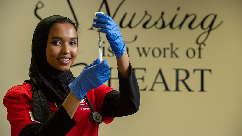 Nursing student wearing gloves and holding a syringe in front of a wall displaying the phrase 'Nursing is a work of heart'