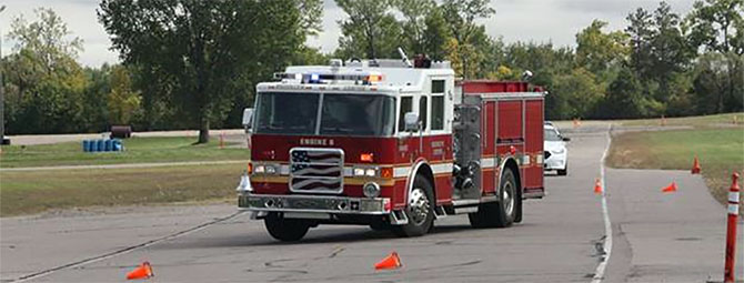 Red fire truck driving through a cone-marked training course on a paved roadway, with traffic cones arranged along the lane and trees and grassy areas in the background
