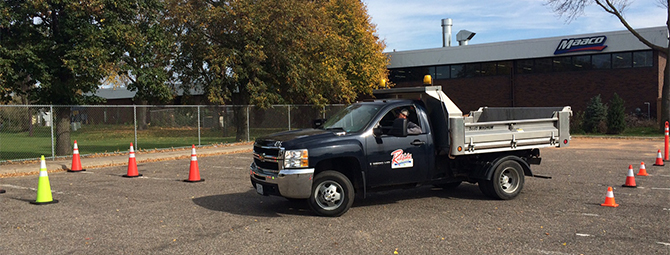 Pickup truck navigating a cone-marked driving course in a parking lot, practicing low-speed maneuvering with an instructor visible in the driver’s seat and a commercial building in the background